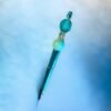 A shimmering teal pen with decorative turquoise and mint green beads on a blue surface.