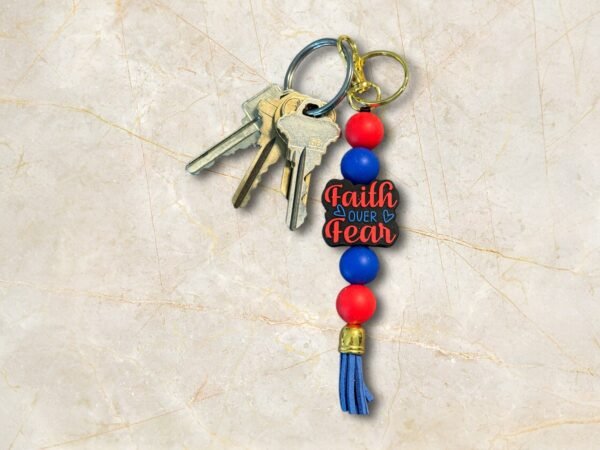 A keychain with keys, red and blue beads, and a charm that says Faith Over Fear on a marble surface.
