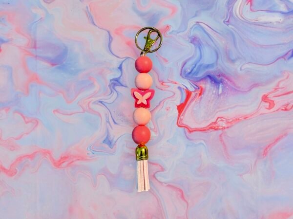 A pink beaded keychain with a butterfly bead, light pink tassel, and gold clasp on a marbled surface.