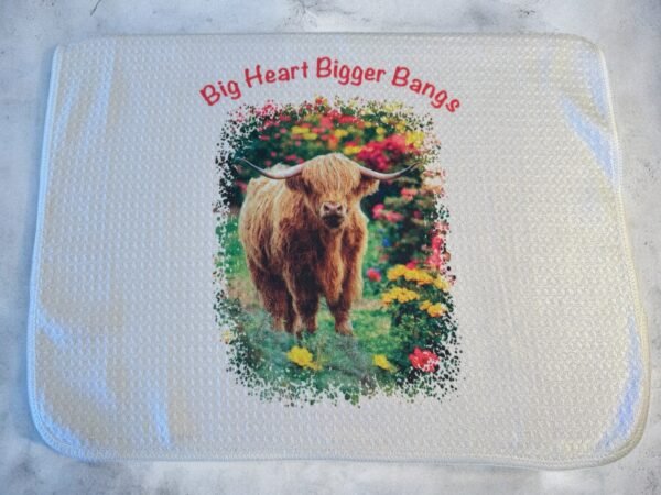 Fluffy Highland cow in colorful flowers on a white dish drying mat with text 'Big Heart Bigger Bangs'.