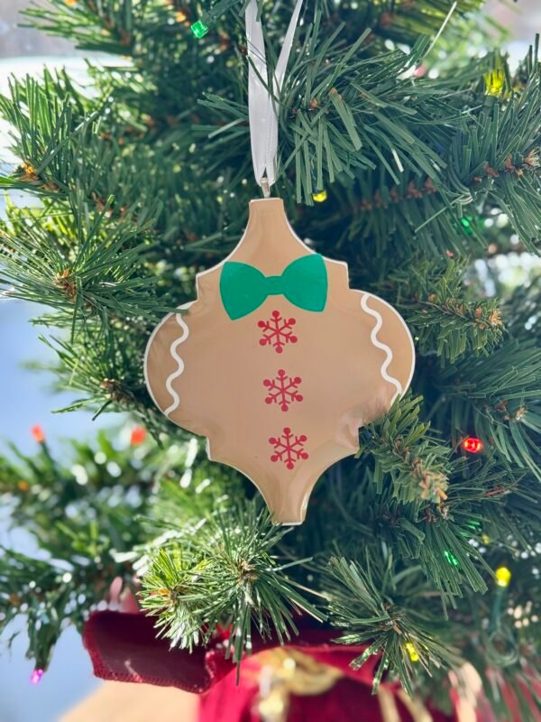 A tan gingerbread man shaped ornament with a green bow tie and red snowflakes hangs on a Christmas tree.