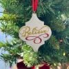 A white ornate Christmas ornament with gold glitter text 'Believe' and red flourish hangs from a red ribbon