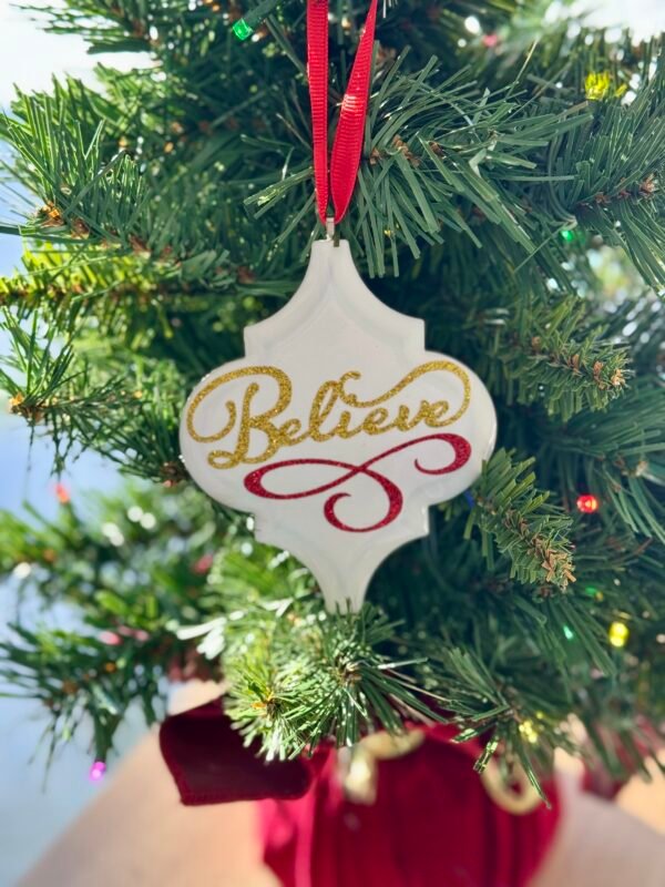 A white ornate Christmas ornament with gold glitter text 'Believe' and red flourish hangs from a red ribbon