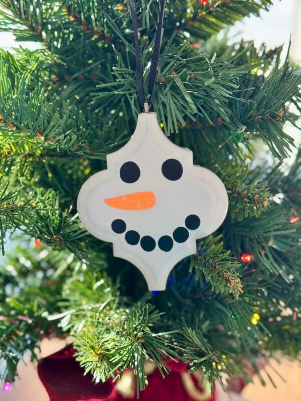 A white snowman ornament with black eyes and a carrot nose hangs on a green Christmas tree.