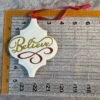 White quatrefoil ornament with gold glitter "Believe" and a red glitter swirl, on a grey ruler.