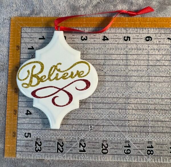 White quatrefoil ornament with gold glitter "Believe" and a red glitter swirl, on a grey ruler.
