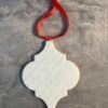A white, blank quatrefoil-shaped ornament with a red ribbon loop on gray fabric.