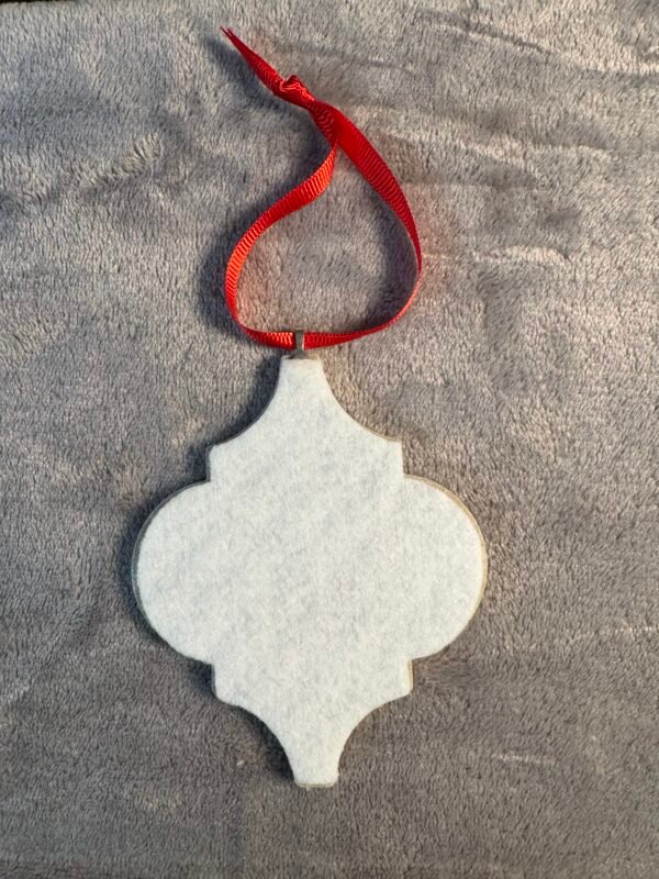 A white, blank quatrefoil-shaped ornament with a red ribbon loop on gray fabric.
