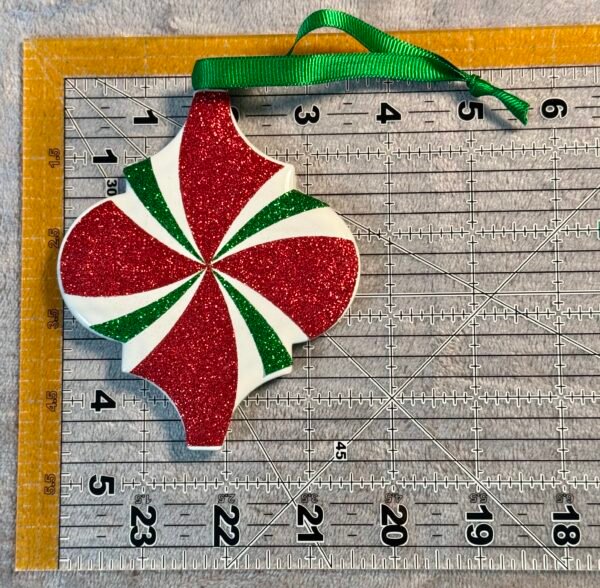 A festive red and green glitter ornament with a white swirled design and a green ribbon.