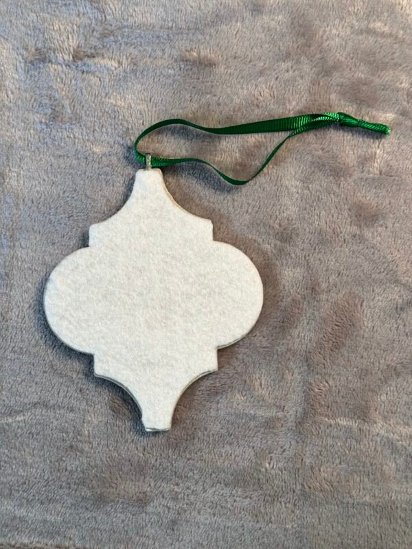 White felt ornament with an arabesque shape and a green ribbon on a gray fuzzy surface.