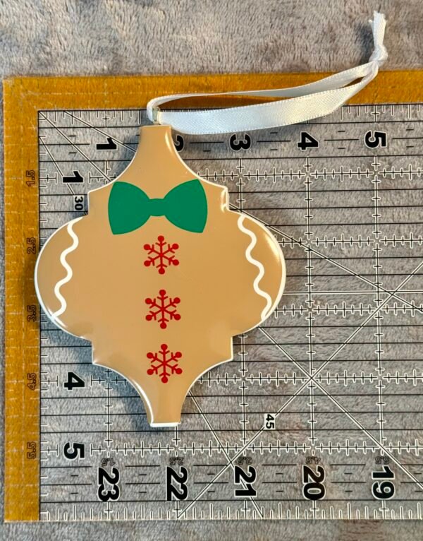 A tan gingerbread man ornament, with a green bow tie and red snowflakes, on a measuring ruler.