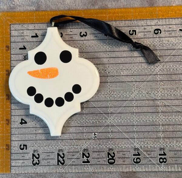 A white snowman-shaped ornament featuring black eyes, an orange glitter nose, and a black ribbon.