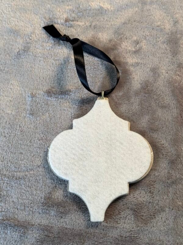 A white quatrefoil-shaped ornament with a black ribbon tie, resting on a gray fuzzy fabric surface.