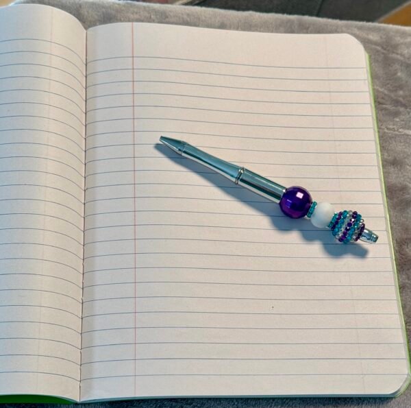 A decorative silver pen with purple and turquoise beads rests on an open lined notebook.