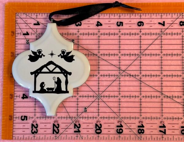 A white ornament featuring a black nativity scene silhouette with Mary, Joseph, baby Jesus, angels