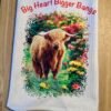 White towel with a shaggy Highland cow among colorful flowers and red 'Big Heart Bigger Bangs' text.