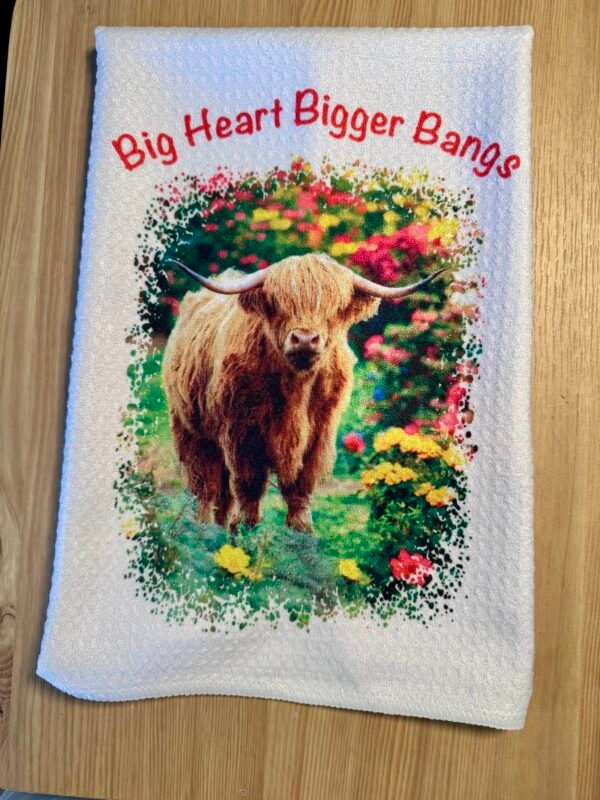White towel with a shaggy Highland cow among colorful flowers and red 'Big Heart Bigger Bangs' text.