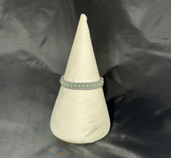A beaded bracelet with green and silver beads, displayed on a cream conical stand.