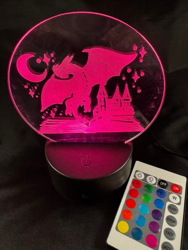 A glowing pink acrylic lamp features an etched design of a dragon, castle, open book, crescent moon