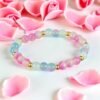 Pastel beaded stretch bracelet with pink and light blue round beads separated by small gold spacer beads, arranged in a circle and displayed on a white surface surrounded by soft pink rose petals.