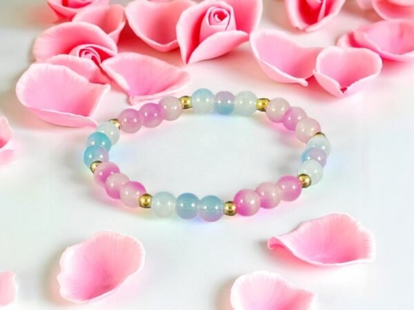 Pastel beaded stretch bracelet with pink and light blue round beads separated by small gold spacer beads, arranged in a circle and displayed on a white surface surrounded by soft pink rose petals.