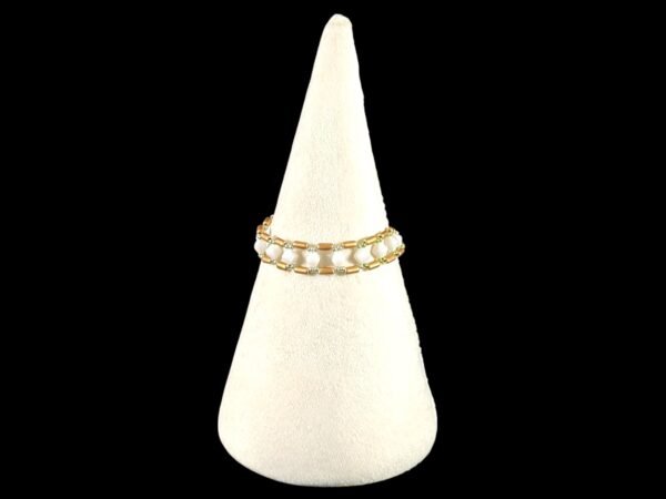 A gold and white beaded ring with small green accents, displayed on a cream velvet cone.