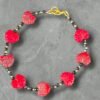 Black beaded bracelet featuring red carved leaf-shaped beads spaced evenly around the strand, accented with small gold and black spacer beads and finished with a gold lobster clasp, displayed on a gray stone background.